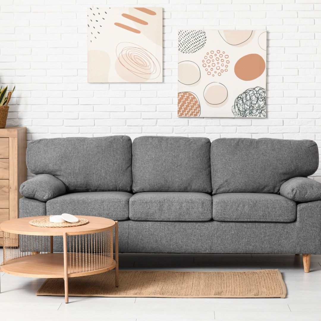 Ein modernes Wohnzimmer mit einem grauen Sofa, einem runden Holztisch und zwei abstrakten Gemälden an einer weißen Ziegelwand.