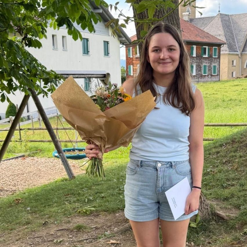 Eine junge Frau steht auf einer Rasenfläche und hält einen Blumenstrauß und einen weißen Umschlag in der Hand. Sie lächelt und scheint für ein Foto zu posieren. Hinter ihr steht ein Baum, ein Haus und eine Schaukel.