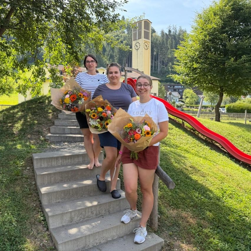 Drei Frauen stehen auf Betonstufen und halten Blumensträuße. Sie lächeln und scheinen für ein Foto zu posieren. Hinter ihnen befindet sich eine Rutsche und ein Kirchturm.