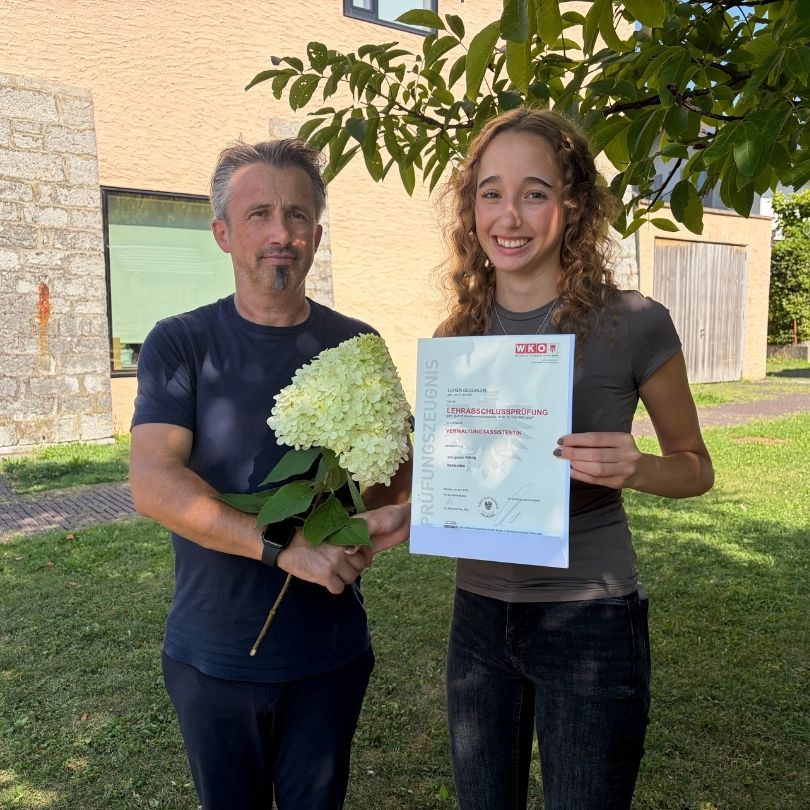 Ein Mann und eine Frau stehen vor einem Gebäude, die Frau hält ein Zertifikat und einen Blumenstrauß, beide lächeln.