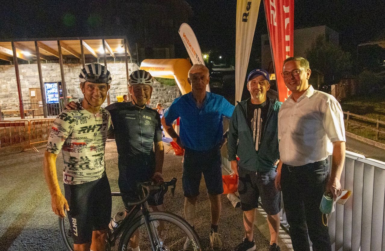Five men stand together at night, two with bicycles, one in a blue shirt, one in a green jacket, and one in a white shirt. Behind them, there is a building with a lit-up sign.