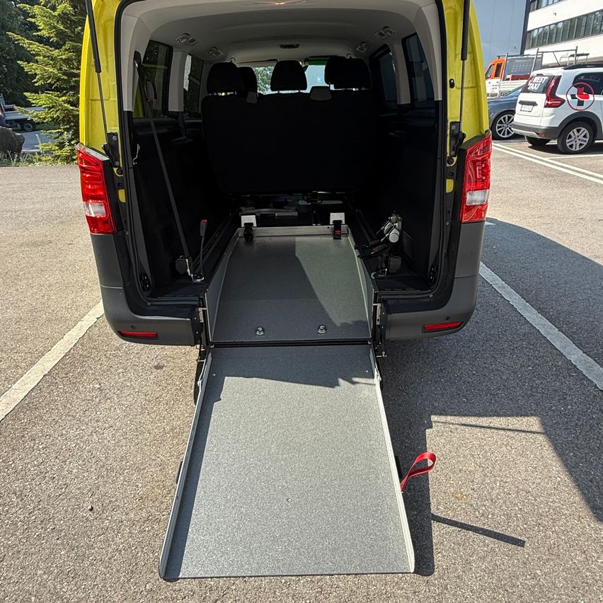The back of a yellow and gray van with its ramp extended, parked in a parking lot with trees and other vehicles in the background.