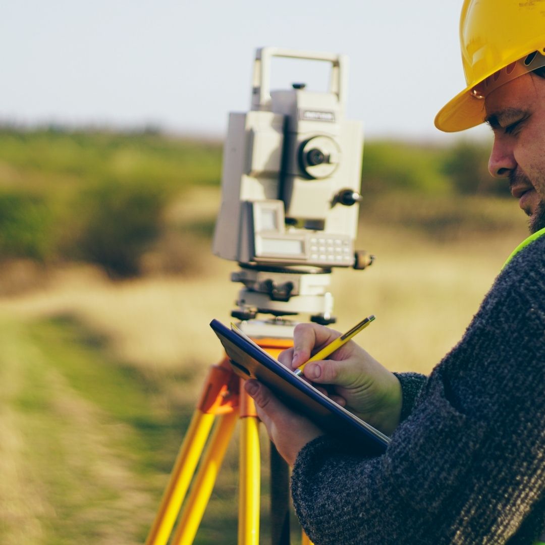 A man in a yellow hard hat is writing on a tablet while using a surveying instrument in a field.