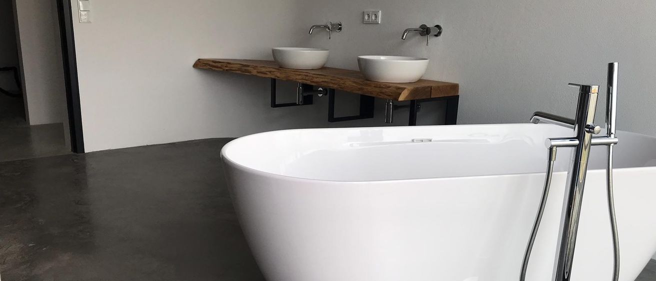 Ein modernes Badezimmer mit einer weißen Badewanne, zwei weißen Waschbecken auf einem Holzregal und einem Duschkopf auf einem Ständer. Das Badezimmer hat weiße Wände und grauen Betonboden.