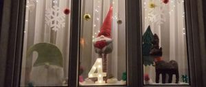 Ein Fensterdisplay mit Weihnachtsdekorationen. Es zeigt einen Weihnachtsmannhut, die Zahl 4 und einen Rentier. Schneeflocken und Weihnachtskugeln hängen am Fensterrahmen. Der Hintergrund ist weiß.