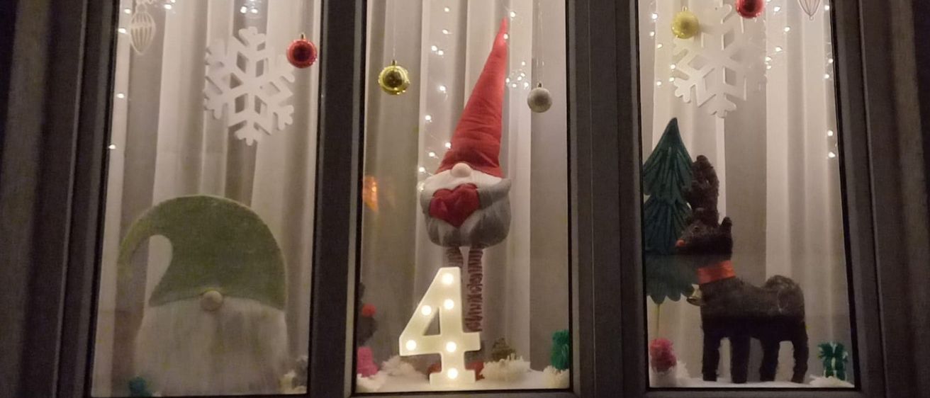 Ein Fensterdisplay mit Weihnachtsdekorationen. Es zeigt einen Weihnachtsmannhut, die Zahl 4 und einen Rentier. Schneeflocken und Weihnachtskugeln hängen am Fensterrahmen. Der Hintergrund ist weiß.