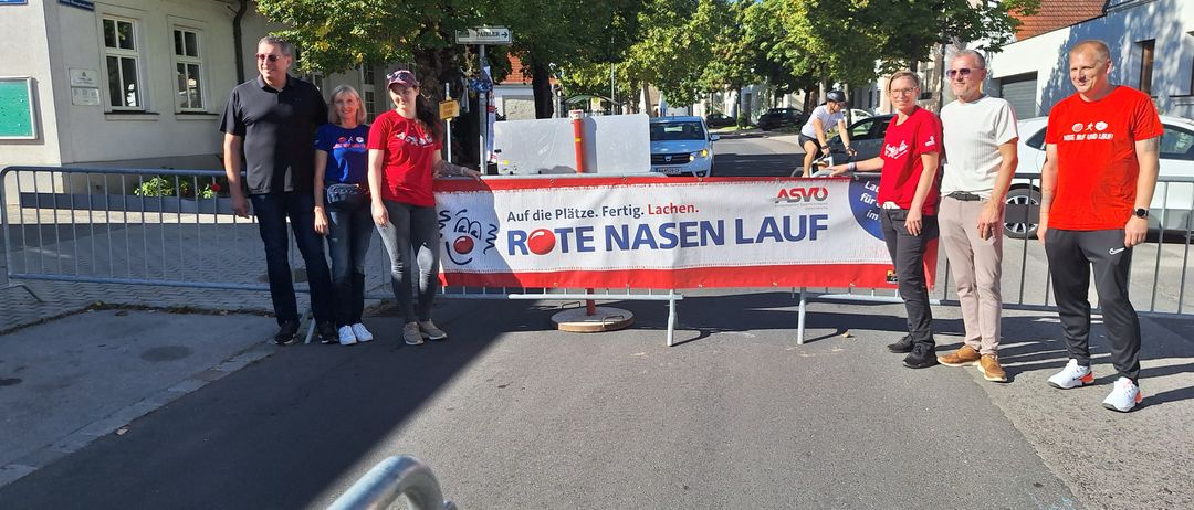 Drei Frauen stehen hinter einem Banner mit der Aufschrift 'Rote Nasen Lauf' mit einem Radfahrer im Hintergrund.