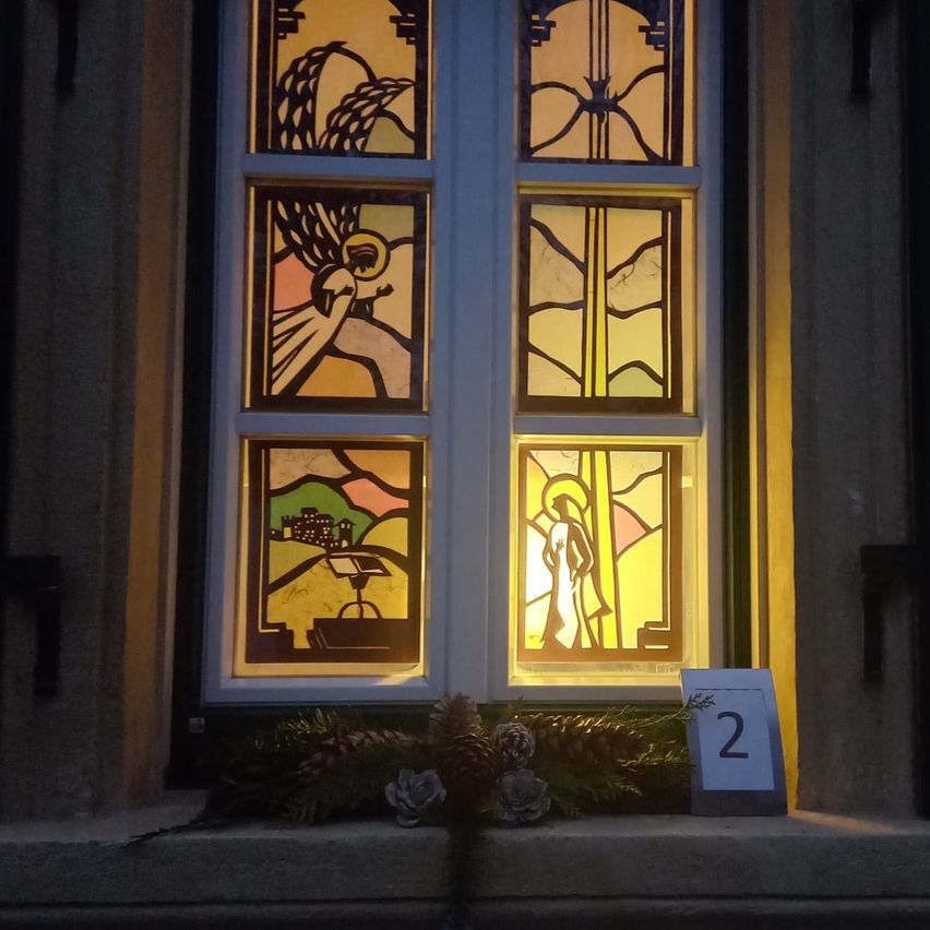 Ein Buntglasfenster zeigt einen Engel, eine Person und eine Landschaft. Es ist von hinten beleuchtet. Das Fenster ist mit Tannenzapfen und Blumen geschmückt. Eine Nummer 2 befindet sich neben dem Fenster.