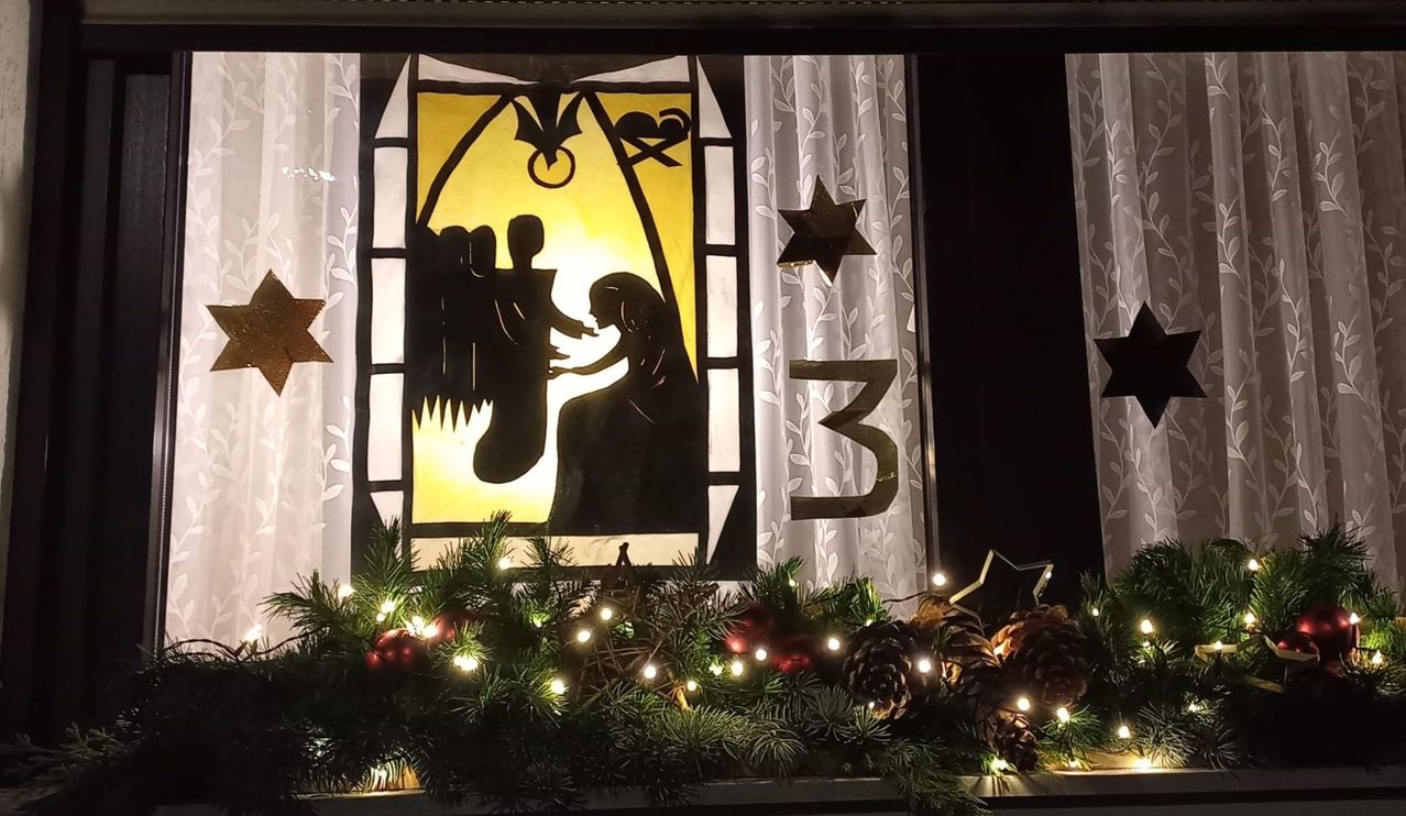 A Christmas window decoration with a nativity scene and stars. The window has a Christmas wreath with lights.