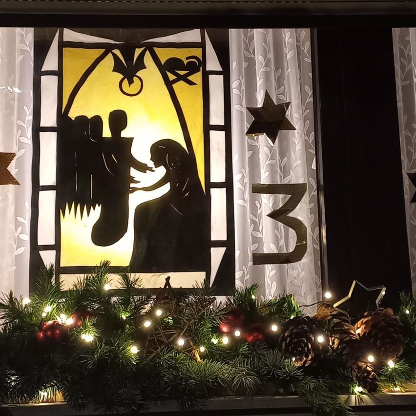 Ein Weihnachtsfensterschmuck mit einer Krippenszene und Sternen. Das Fenster hat eine Weihnachtsgirlande mit Lichtern.