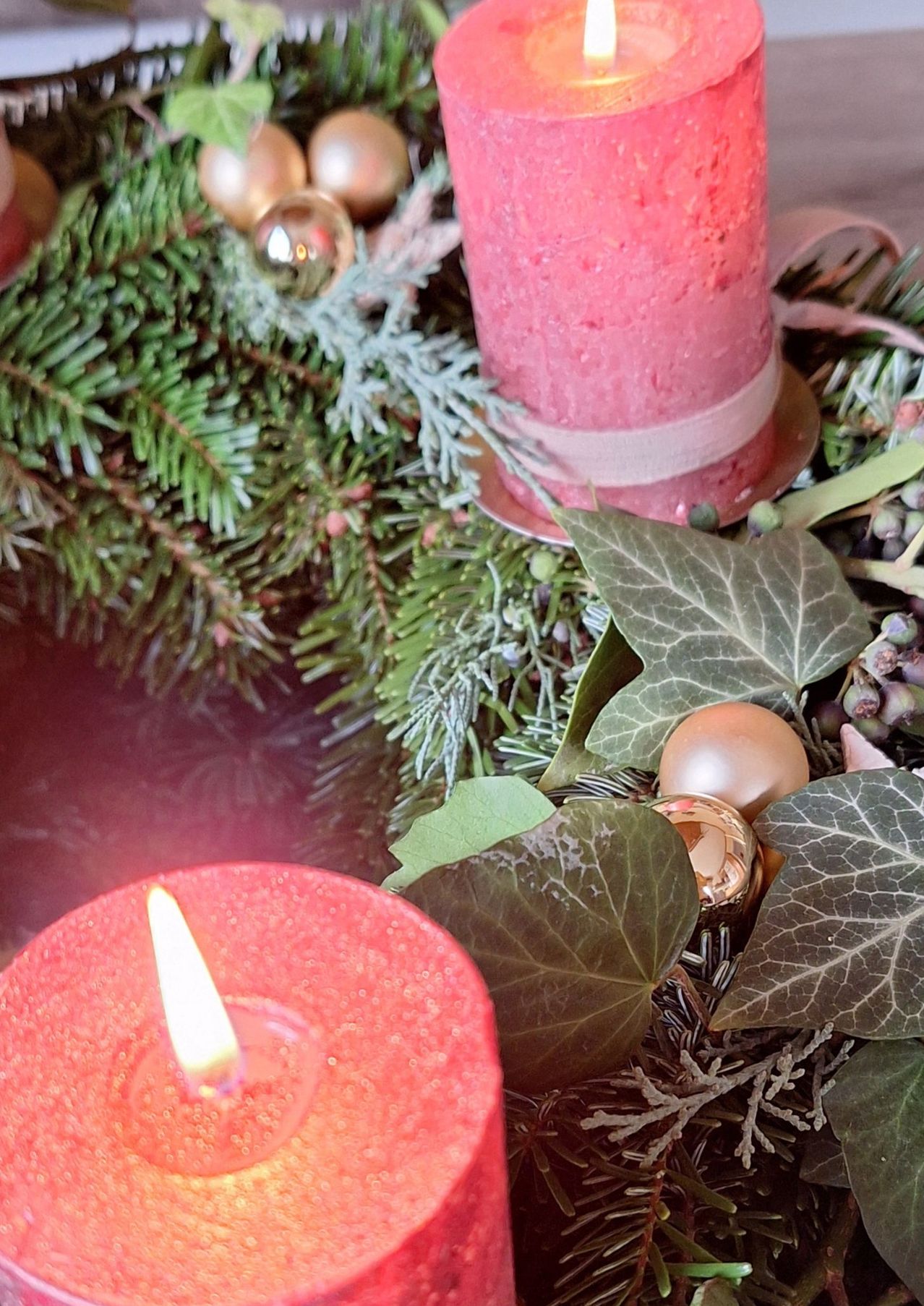 Ein Weihnachtskranz mit zwei roten Kerzen, eine leuchtet, umgeben von Blättern und Tannenzapfen, mit goldenen Ornamenten.