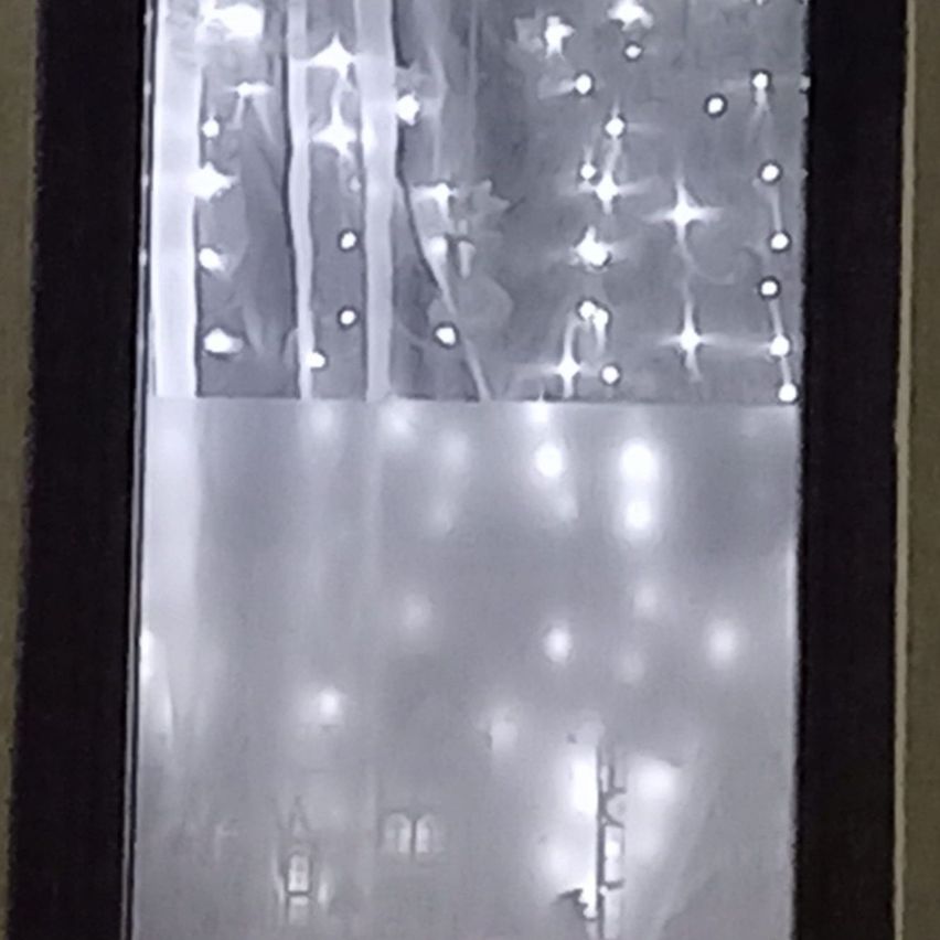 A window with a curtain, showing illuminated lights in a decorative pattern, resembling stars, with a blurred background.