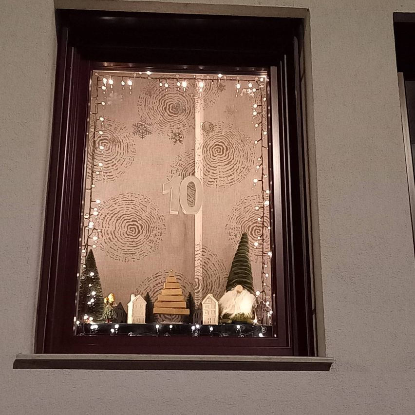 Ein Fenster zeigt eine Weihnachtsszene mit der Nummer 10, einem Gnom und einem Lebkuchenhaus, umgeben von Lichtern und Schneeflocken auf der Scheibe.