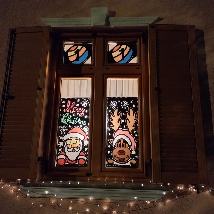 Ein Fenster mit braunen Fensterläden zeigt zwei dekorierte Glasscheiben. Eine zeigt den Weihnachtsmann und die andere ein Rentier, beide mit Weihnachtsgrüßen. Das Fenster ist mit Weihnachtslichtern geschmückt.