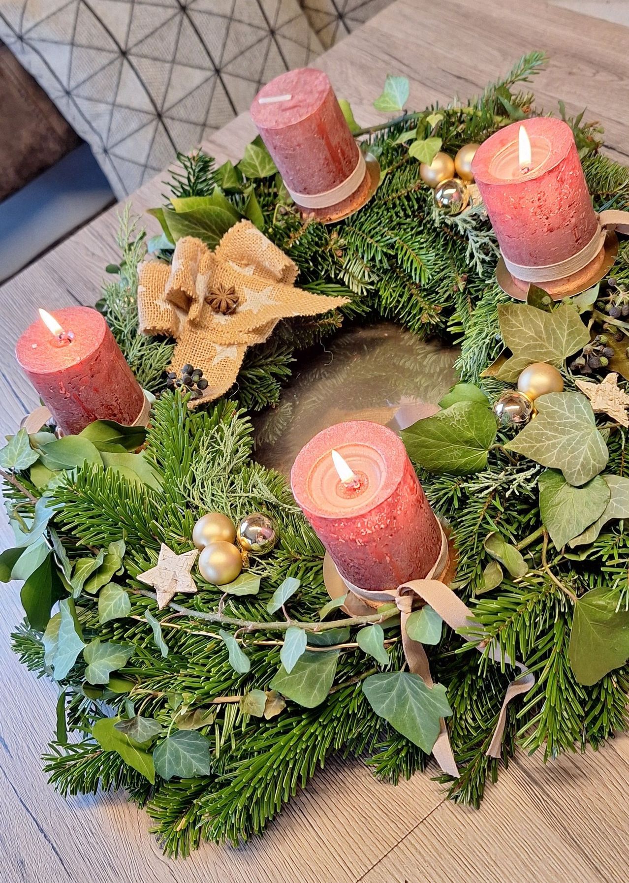 Ein Kranz mit vier brennenden Kerzen, in Burlap gewickelt, geschmückt mit grünen Blättern, goldenen Ornamenten und Sternen, auf einer Holzoberfläche ausgestellt.