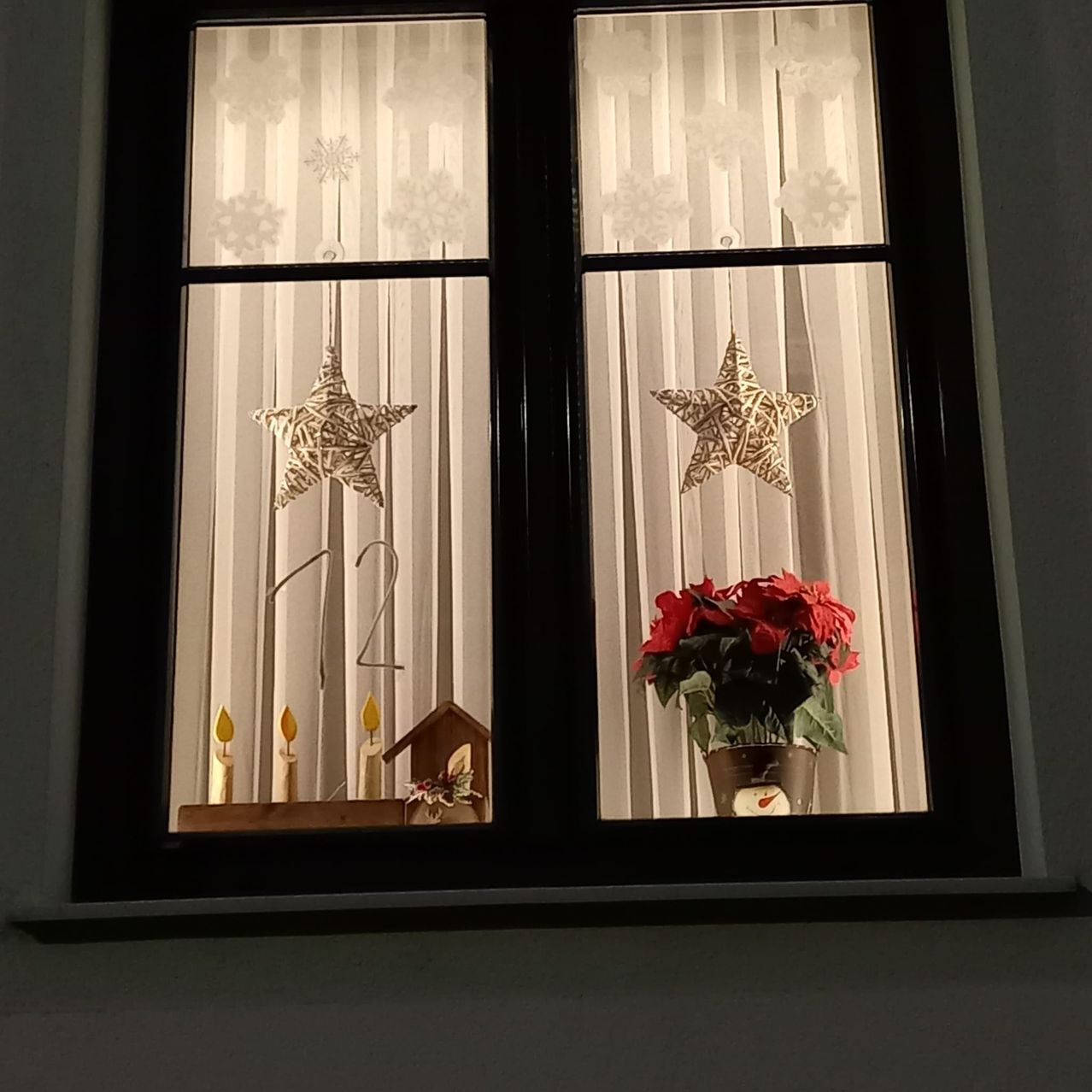 Ein Fenster mit zwei dekorativen Sternen, Schneeflocken und einer Kerzenanzeige. Eine rote Blume in einem Topf ist auf der rechten Seite.