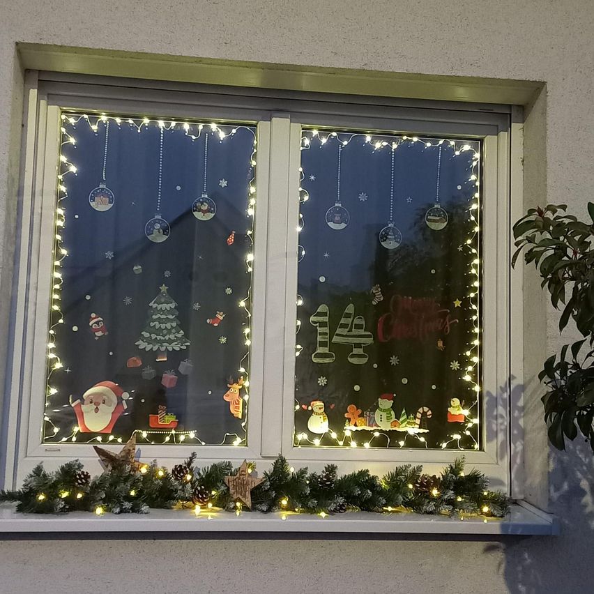 Ein Fenster mit Weihnachtsdekorationen, einschließlich einer Figur von Santa Claus, einem Weihnachtsbaum und Schneeflocken. Der Fenstersims hat eine dekorative Girlande mit Tannenzapfen und Lichtern.