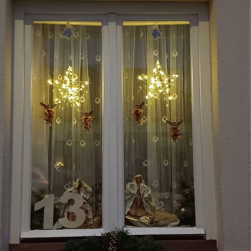 Eine Fensterdekoration zeigt zwei leuchtende Sterndekorationen mit Engeln darüber und einer Krippe darunter, eine Seite mit der Nummer 13 und die andere mit einer Frau und einem Kind.