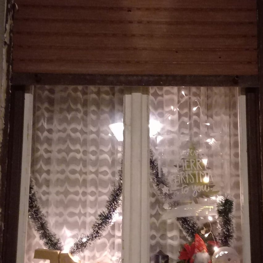 Ein Fenster, geschmückt mit einem Weihnachtskranz und einem dekorativen Schild mit der Aufschrift 'Frohe Weihnachten an Sie'. Der Kranz enthält Lichter, und das Fenster ist teilweise mit weißen Vorhängen bedeckt.