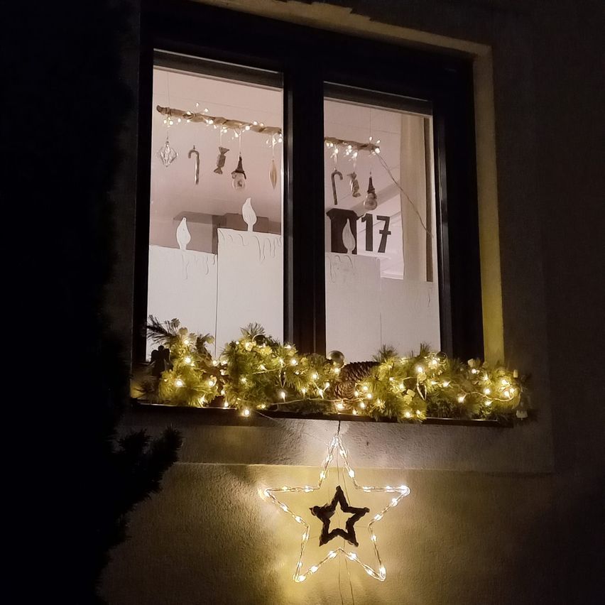 Ein Fensterdisplay mit Weihnachtsdekorationen, einschließlich einer Girlande mit Lichtern, Ornamenten und einem Stern auf der Fensterbank. Das Fenster zeigt Reflexionen von Lichtern und Ornamenten im Inneren.