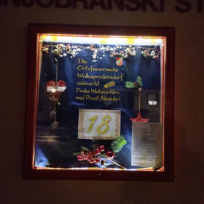 Ein gerahmtes Display mit blauem Hintergrund, beleuchtet mit gelben Lichtern. Es zeigt eine dekorative Banner mit 'Die Ortsfeuerwehr Wulkaprodersdorf wünscht Frohe Weihnachten und Prosit Neujahr!' in Gelb, einem herzförmigen Ornament und einem roten Stern. Die Zahl '18' ist ebenfalls zu sehen. Ein Papier mit Text ist auf der rechten Seite platziert.