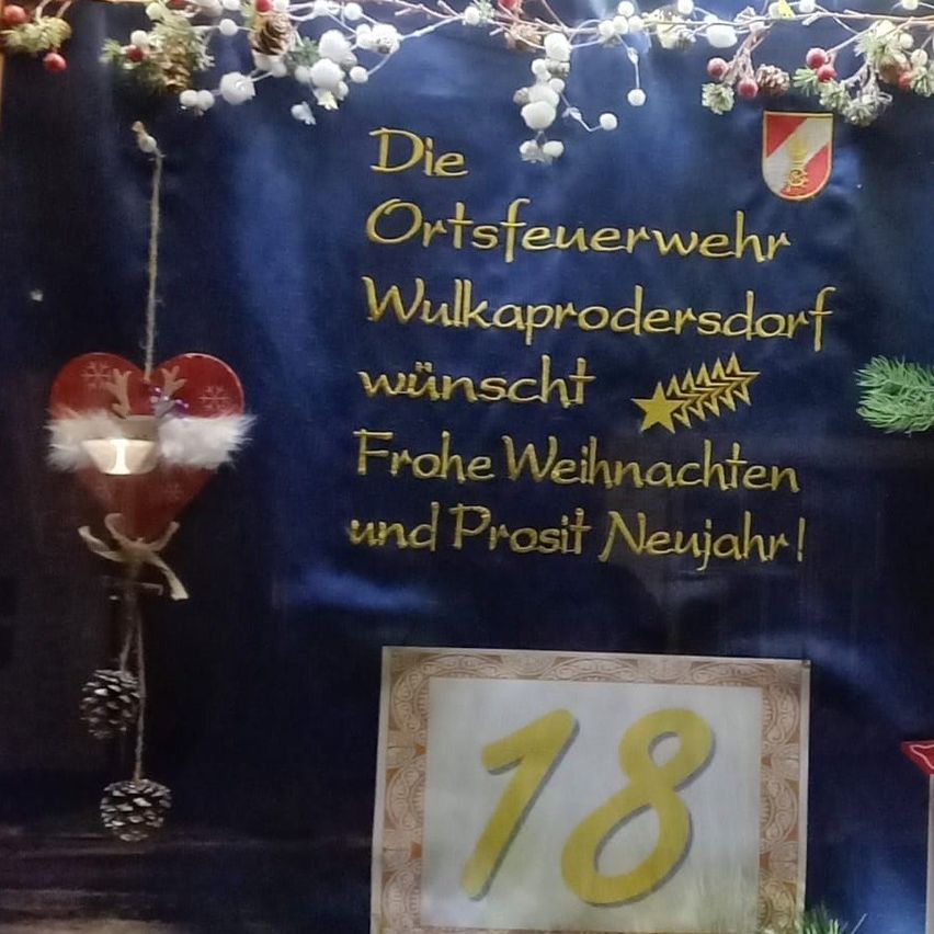 Ein blauer Banner mit Weihnachtsdekorationen zeigt eine Botschaft der örtlichen Feuerwehr von Wulkaprodersdorf, die allen ein frohes Weihnachtsfest und ein glückliches neues Jahr wünscht. Die Nummer 18 ist prominent in Gelb angezeigt.