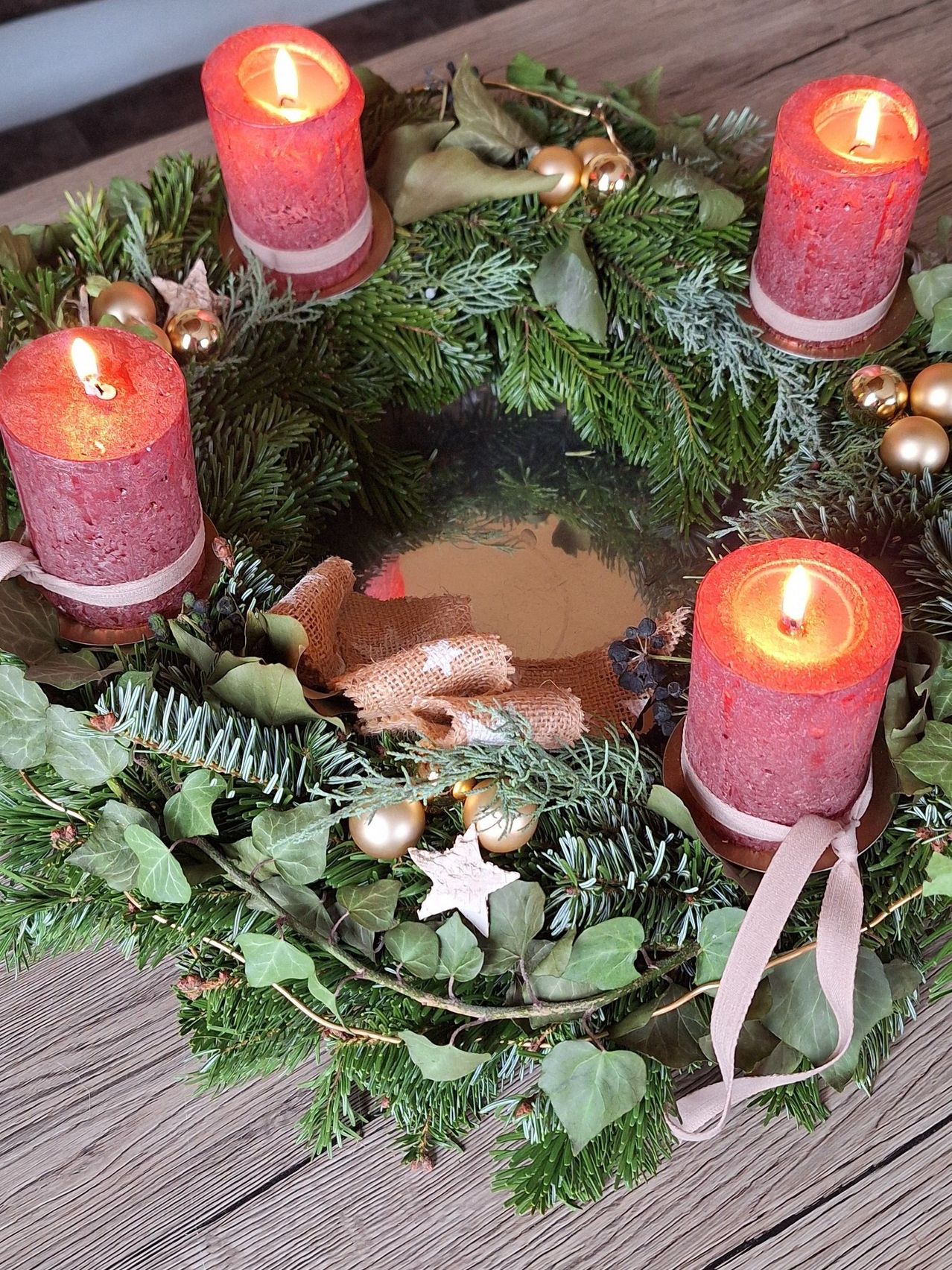 Ein Kranz mit vier brennenden Kerzen, umgeben von Grün, Tannennadeln und goldenen Ornamenten. Die Kerzen sind in Juteseilen eingewickelt.