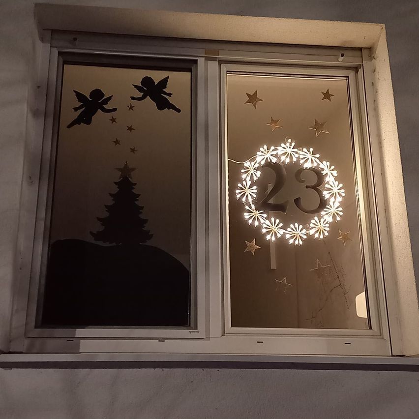 Ein Fensterschmuck mit einem beleuchteten Kranz, der die Nummer 23 zeigt, mit Engelssilhouetten und einem Weihnachtsbaum im Hintergrund.