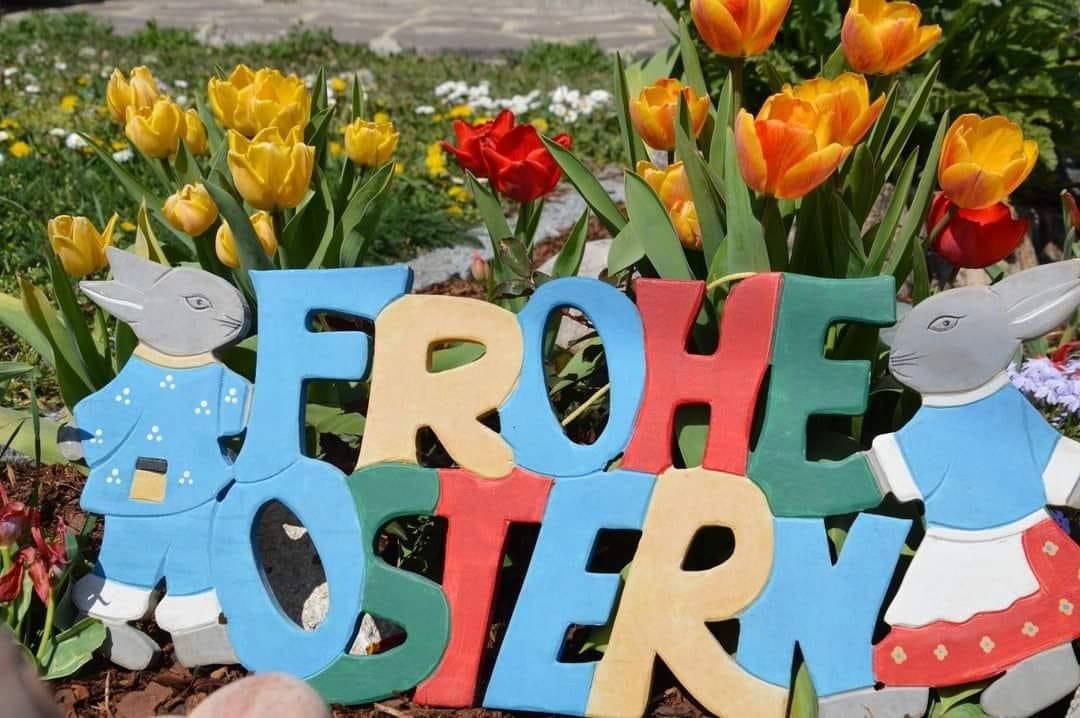Colorful letters spell Frohe Ostern surrounded by tulips and garden figurines. The sign sits in a garden with green grass and rocks.