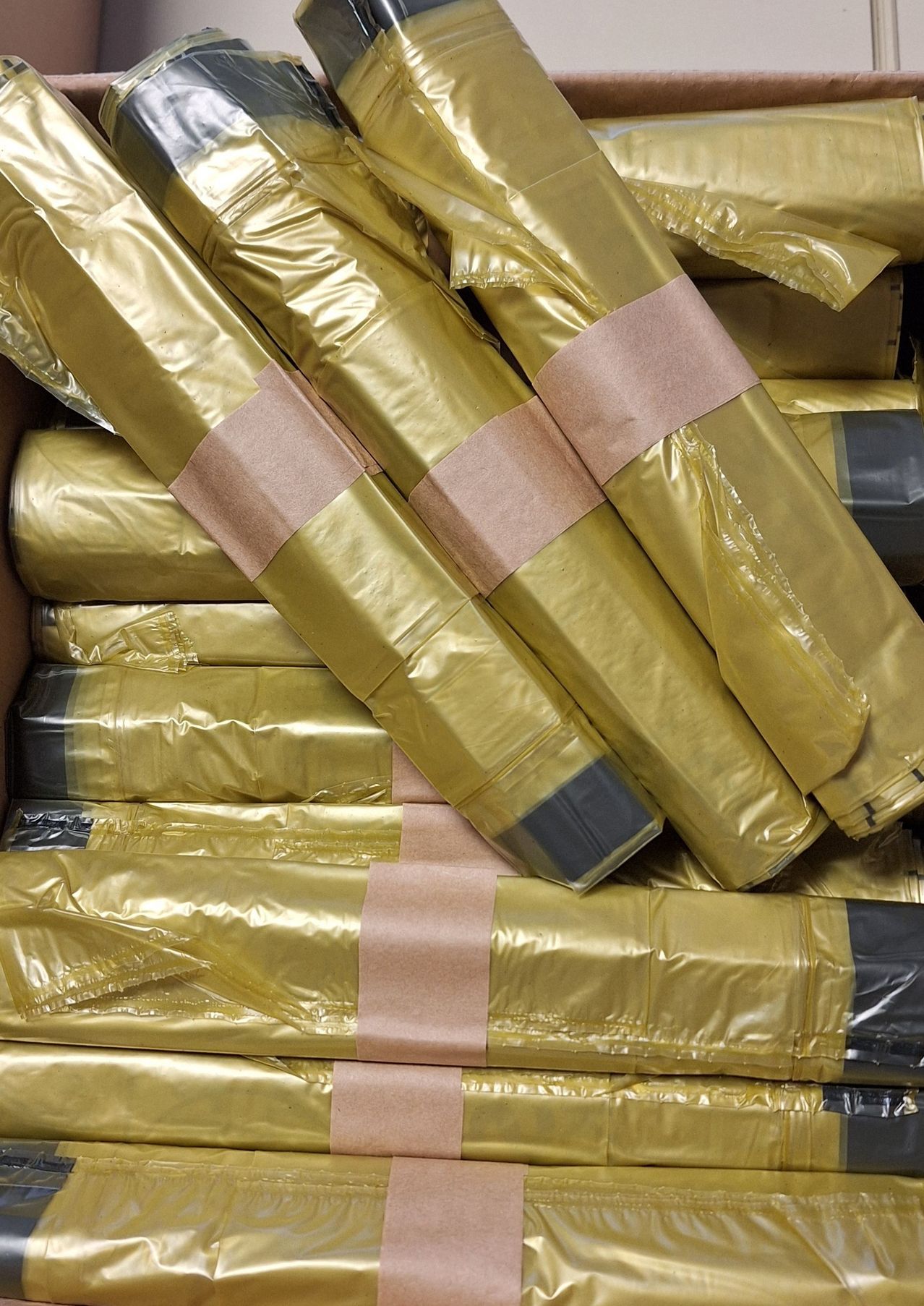 A stack of golden-wrapped packages secured with brown tape. The packages are in a box, stacked neatly on top of each other. Each package has a small piece of gray tape at the end.