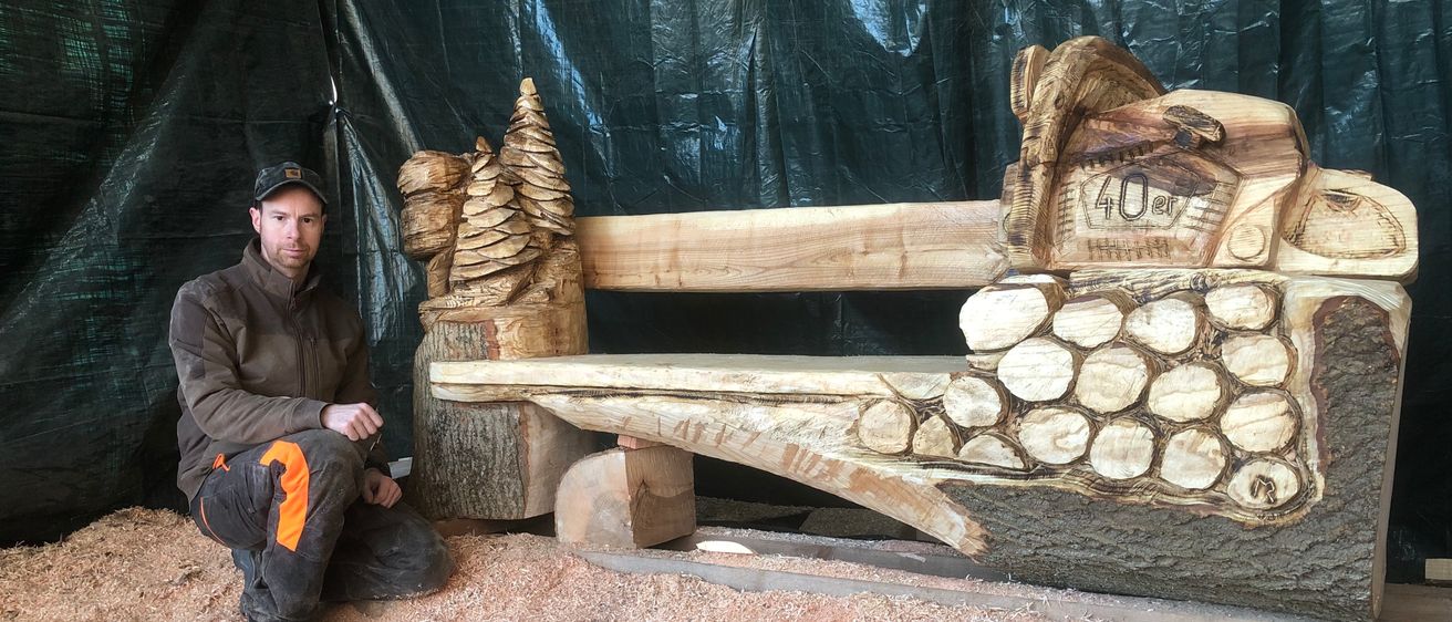 Ein Mann sitzt auf dem Boden neben einer aus einem Baumstamm geschnitzten Bank, mit einem schwarzen Hintergrund hinter ihm.