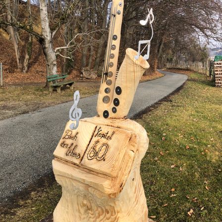 Eine Holzskulptur eines Saxophons mit Noten und einem Buch mit dem Text 'Buch des Lebens 60' auf einem Grasweg.