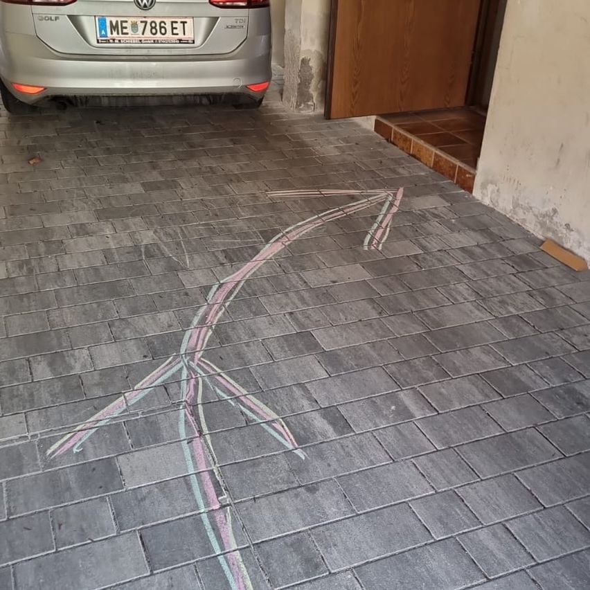 Ein silbernes Auto mit dem Kennzeichen ME 786 ET ist in einer Einfahrt geparkt, mit einer Kreidezeichnung eines Pfades, der zu einer Holztür führt.