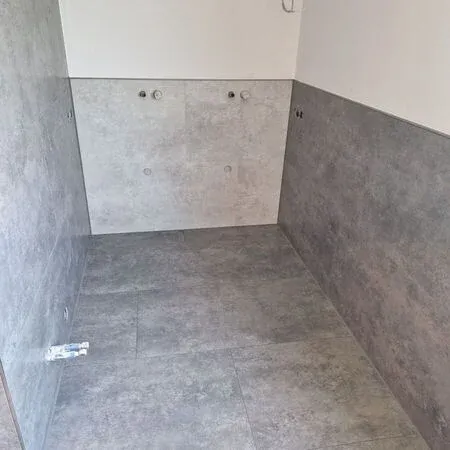 Ein leerer Raum mit grauen gefliesten Böden und Wänden. Drei kreisförmige Löcher sind an der Wand sichtbar.