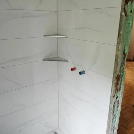 Eine Badezimmerecke mit weißen Marmorwänden und zwei Metallregalen. An der Wand befinden sich zwei Armaturen. Der Boden ist aus Holz.