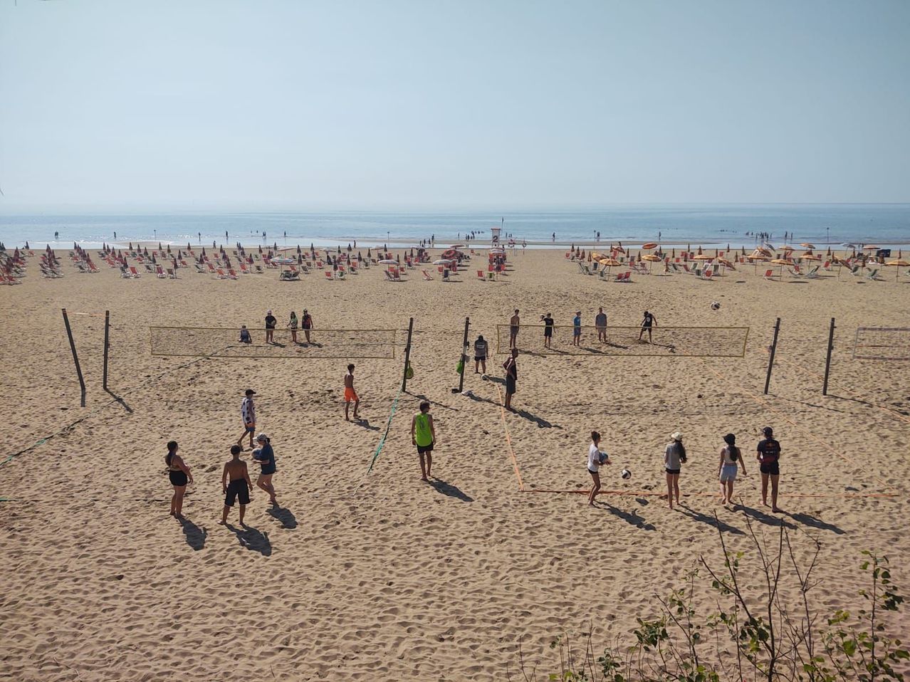 Eine Strandszene mit mehreren Menschen, die Volleyball spielen und sich sonnen, während andere am Strand stehen.