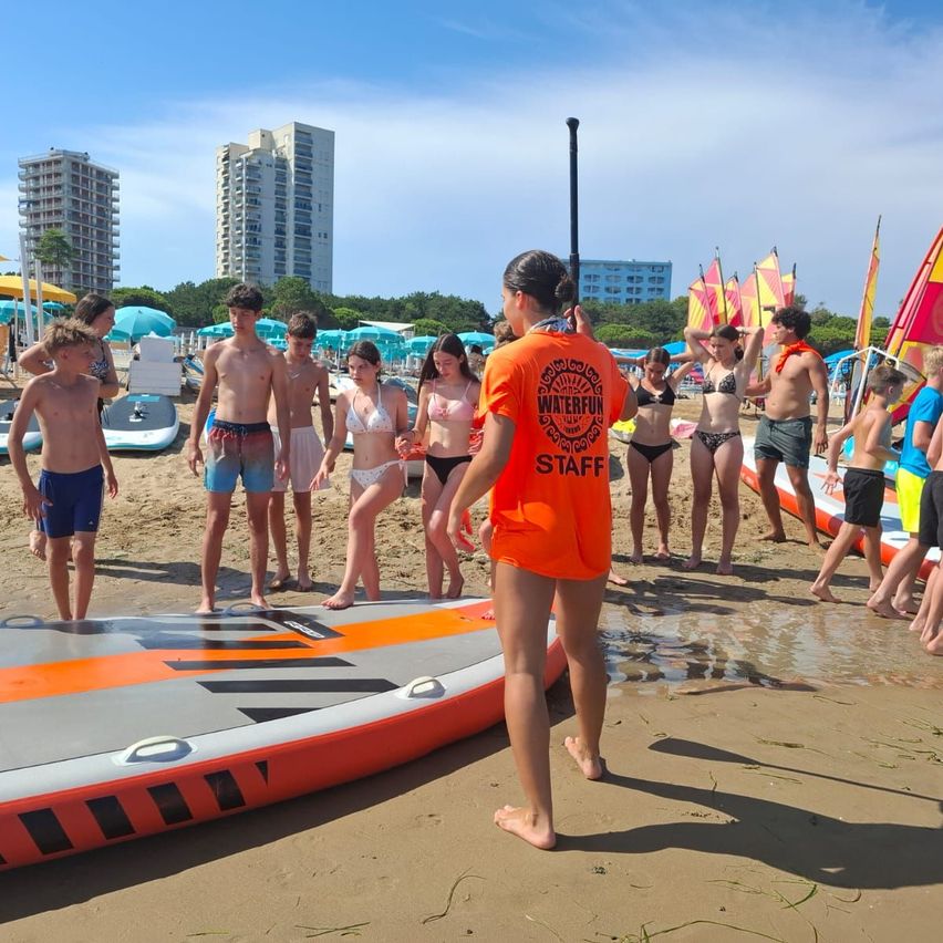 Eine Gruppe von Menschen, wahrscheinlich Touristen, ist am Strand. Eine Frau in einem orangefarbenen Shirt spricht zu ihnen. Mehrere aufblasbare Bretter liegen am Sand.