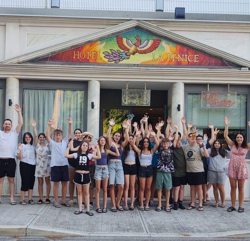 Eine Gruppe von Menschen posiert für ein Foto vor dem Hotel La Fenice. Sie alle lächeln und heben ihre Hände.