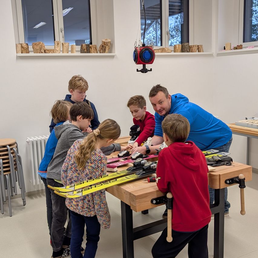 Eine Gruppe von Kindern und einem Mann arbeitet an Skiern auf einer Holzwerkbank in einer Werkstatt.