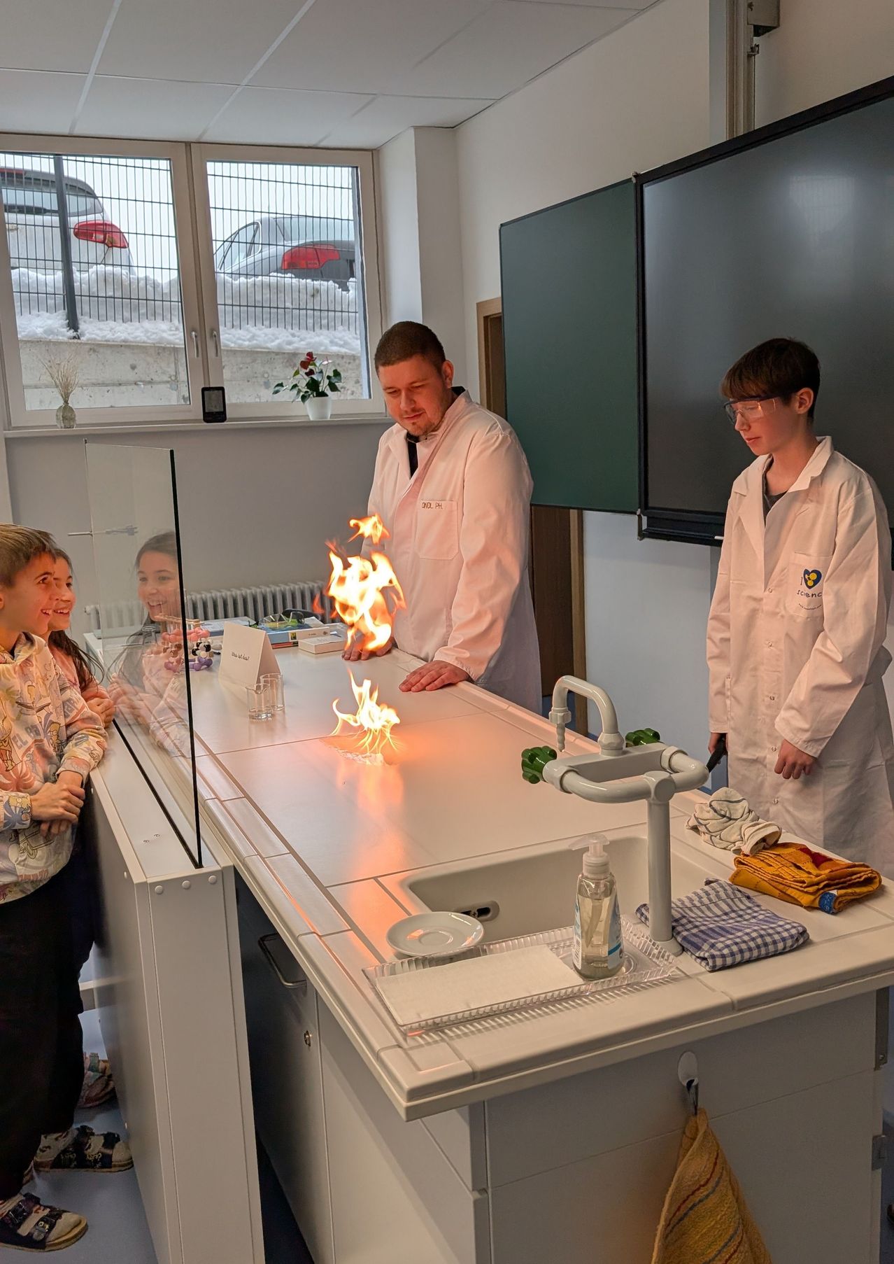 Eine Gruppe von Schülern und einem Lehrer sind in einem Labor. Eine Feuerdemonstration wird mit Sicherheitsvorkehrungen durchgeführt.