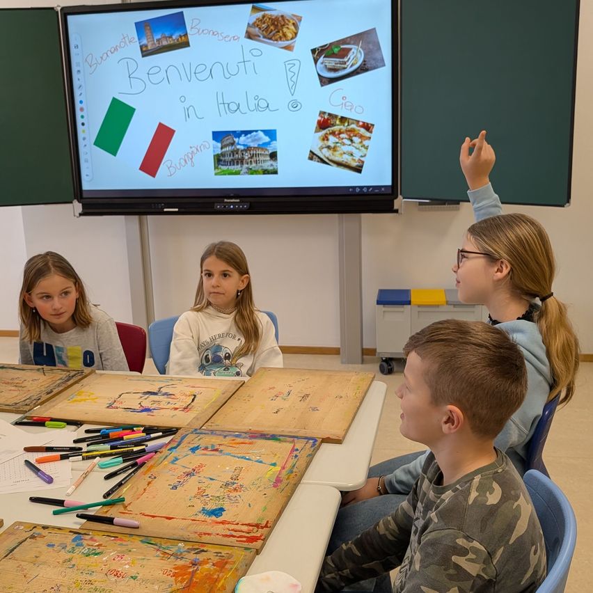 Vier Kinder sitzen in einem Klassenzimmer um einen Tisch herum und zeichnen mit Markern auf Papier. Ein großer Bildschirm zeigt 'Benvenuti in Italia' und Bilder aus Italien. Ein Kind hebt die Hand.