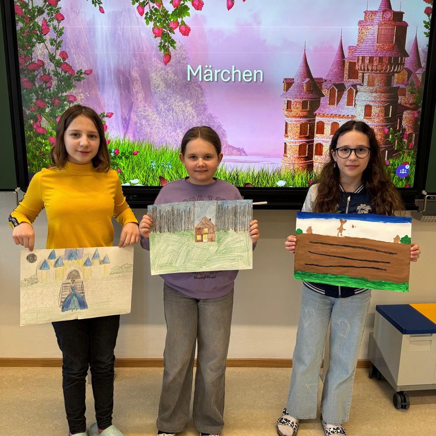Drei junge Mädchen posieren mit ihren Kunstwerken. Sie stehen vor einem Bildschirm, der ein märchenhaftes Bild mit einem Schloss und Blumen zeigt. Jedes Mädchen hält ein Kunstwerk in der Hand.