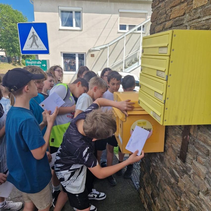 Eine Gruppe von Kindern steht um einen gelben Briefkasten herum. Sie halten Briefumschläge und scheinen etwas zu verschicken. Der Briefkasten ist an einer Steinmauer montiert. Hinter ihnen befindet sich ein Gebäude mit Fenstern und einer Treppe.