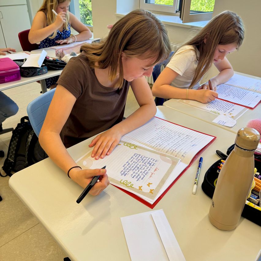 Drei Mädchen sitzen in einem Klassenzimmer und schreiben mit Stiften auf Papier. Eines hat ein braunes Shirt, das andere ein weißes. Bücher und eine Wasserflasche stehen auf dem Tisch.