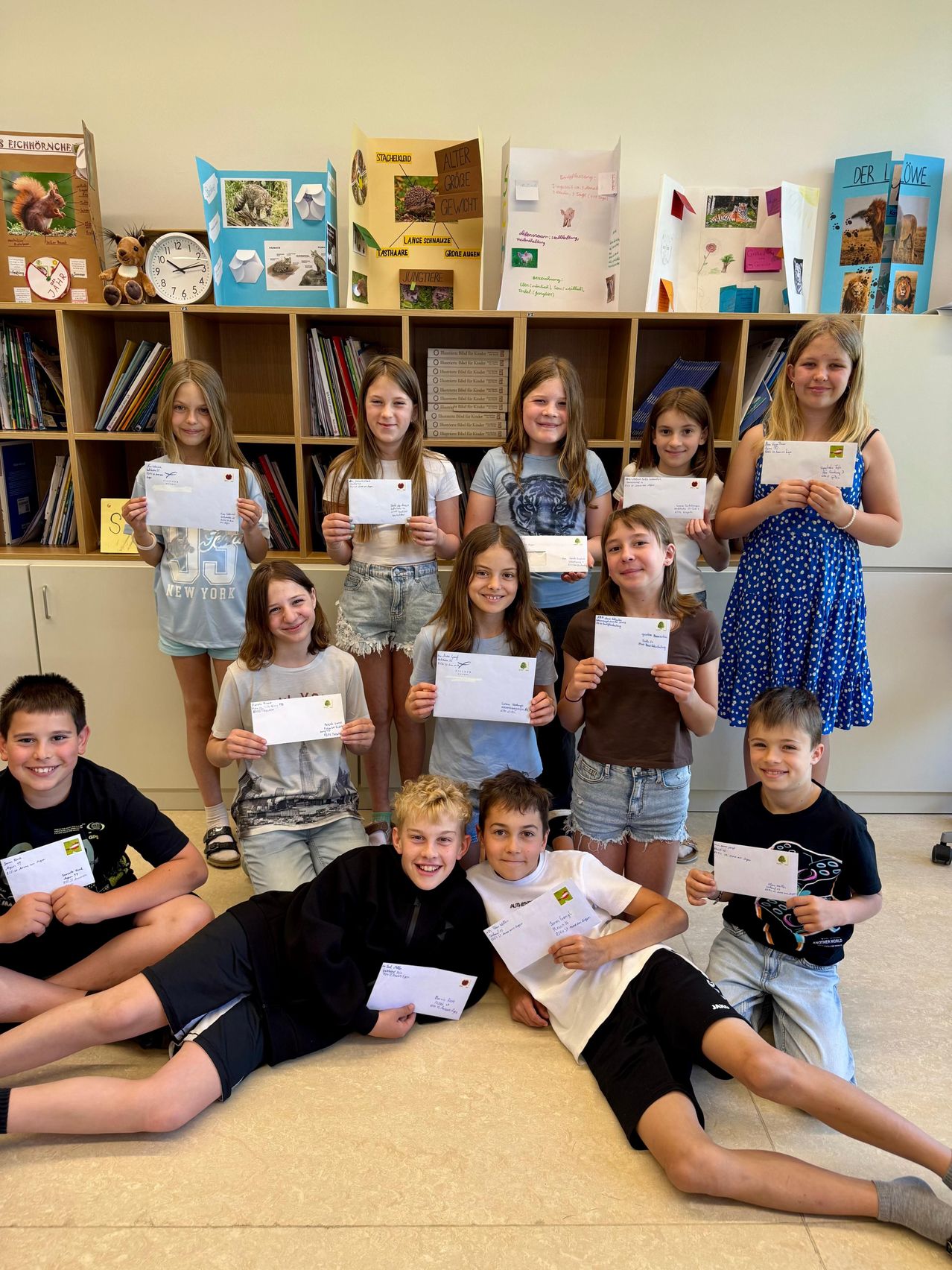 Eine Gruppe von Kindern mit Urkunden, die in einem Klassenzimmer mit Bücherregalen und Dekorationen lächelnd für ein Foto posieren.