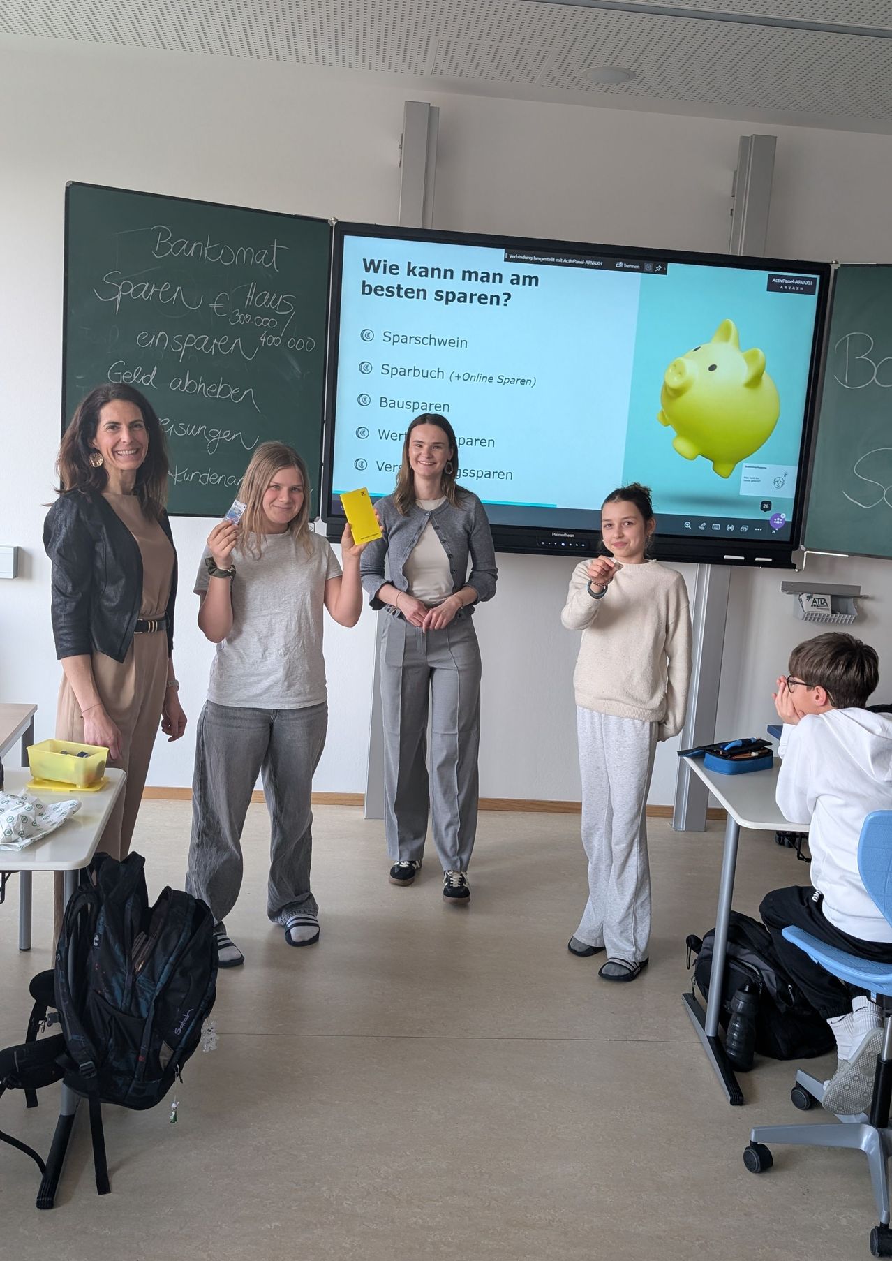 Vier Personen stehen in einem Klassenzimmer und lächeln. Drei von ihnen halten gelbe Objekte, möglicherweise Bücher. Ein großer Bildschirm zeigt ein Sparschwein und Fragen zum Sparen. Ein Junge sitzt an einem Schreibtisch.