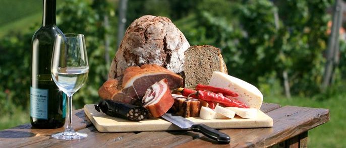 Ein Holztisch im Freien hat eine Flasche Wein, ein Glas und ein Holzschneidebrett mit Brot, Fleisch und Paprika.