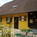 Ein gelbes Haus mit einem braunen Dach, mehreren Fenstern und einer Tür. Ein grüner Tisch mit Stühlen steht davor.