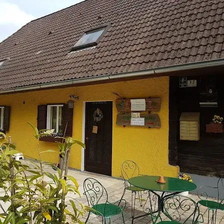 Ein gelbes Haus mit einem braunen Dach, mehreren Fenstern und einer Tür. Ein grüner Tisch mit Stühlen steht davor.