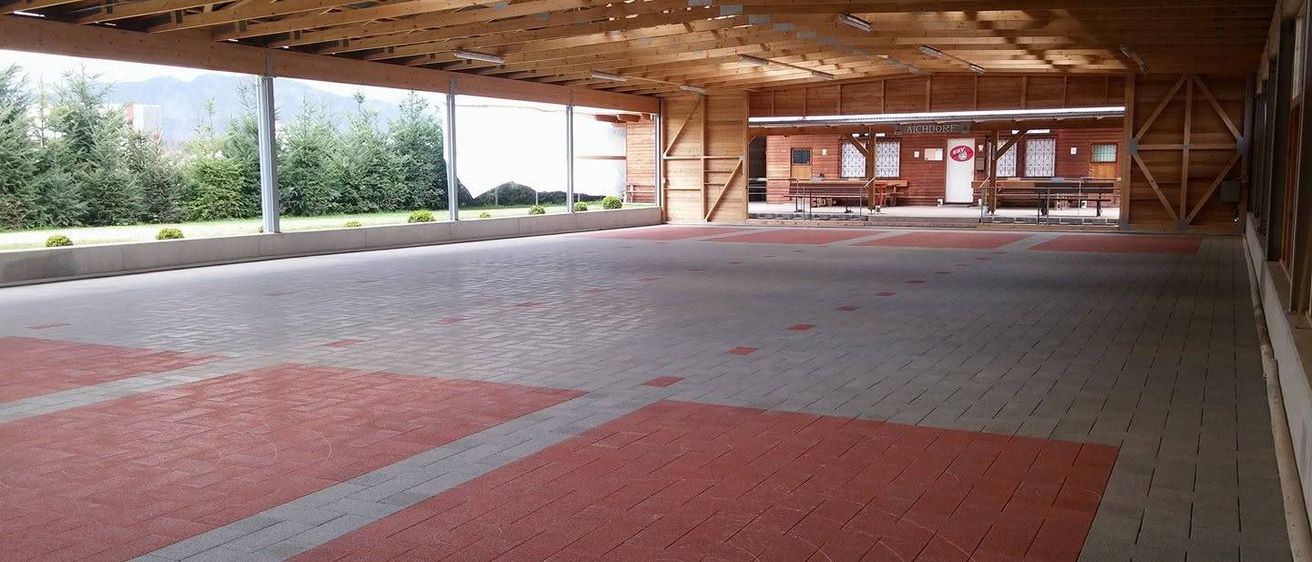 Ein leerer Indoor-Sportbereich mit rotem und grauem Boden, Holzbalken und großen Fenstern.